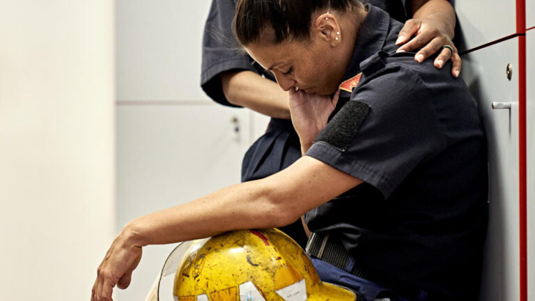 Featured Image – Suicide Concerns- A Guide for Managers Side view of late 30s woman in uniform sitting on bench in fire station locker room with helmet in lap, head down, and eyes closed.